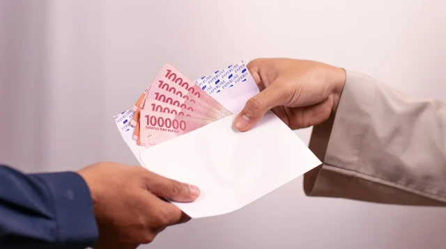 Tangan menyerahkan amplop putih berisi lembaran uang Rp100.000 kepada tangan lain