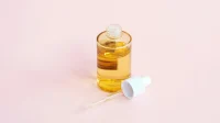 Botol serum kaca bening berisi cairan kuning dengan pipet tergeletak di sampingnya di atas latar merah muda