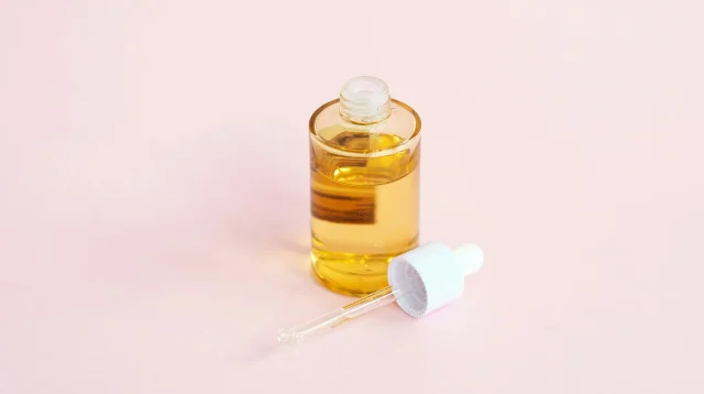 Botol serum kaca bening berisi cairan kuning dengan pipet tergeletak di sampingnya di atas latar merah muda
