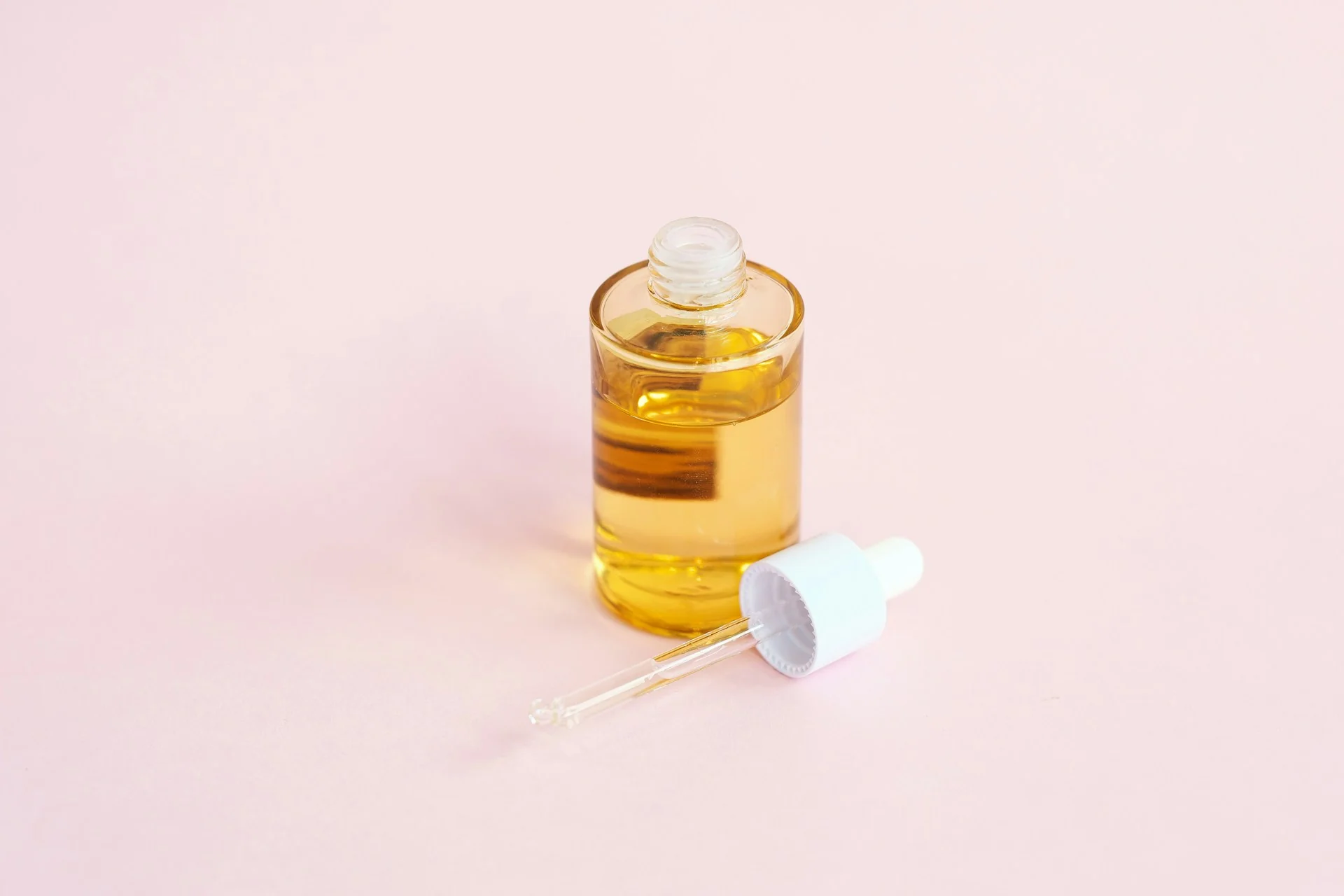 Botol serum kaca bening berisi cairan kuning dengan pipet tergeletak di sampingnya di atas latar merah muda