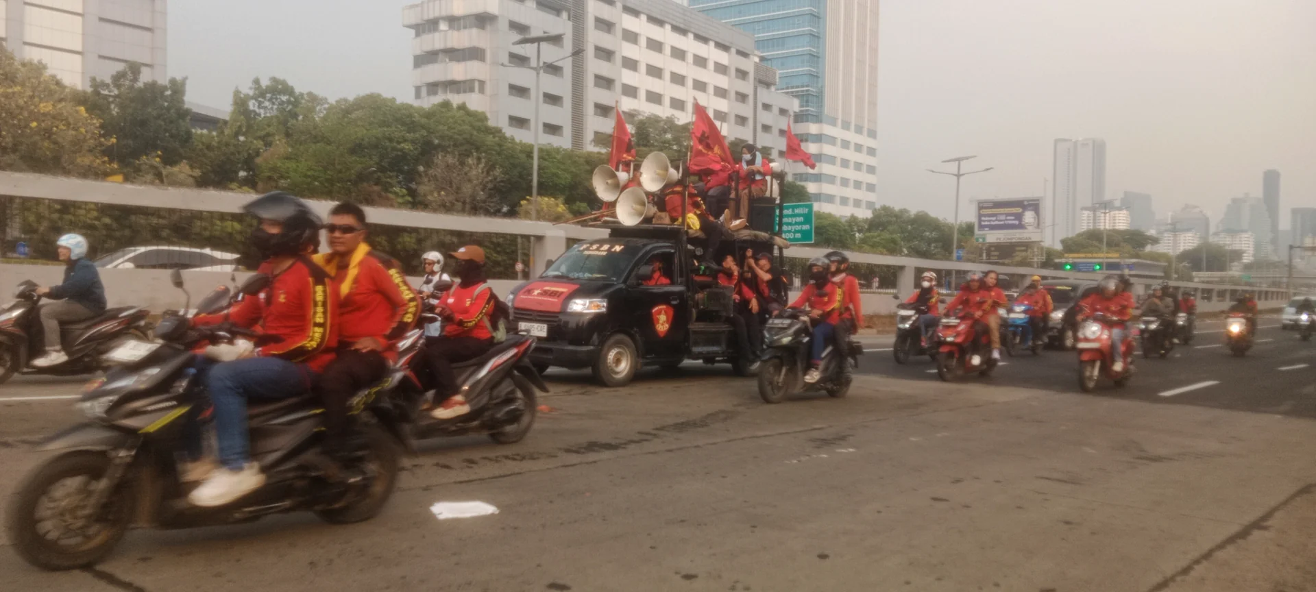 Konvoi buruh berseragam merah naik sepeda motor mengiringi mobil komando berpengeras suara dan bendera di jalan Jakarta