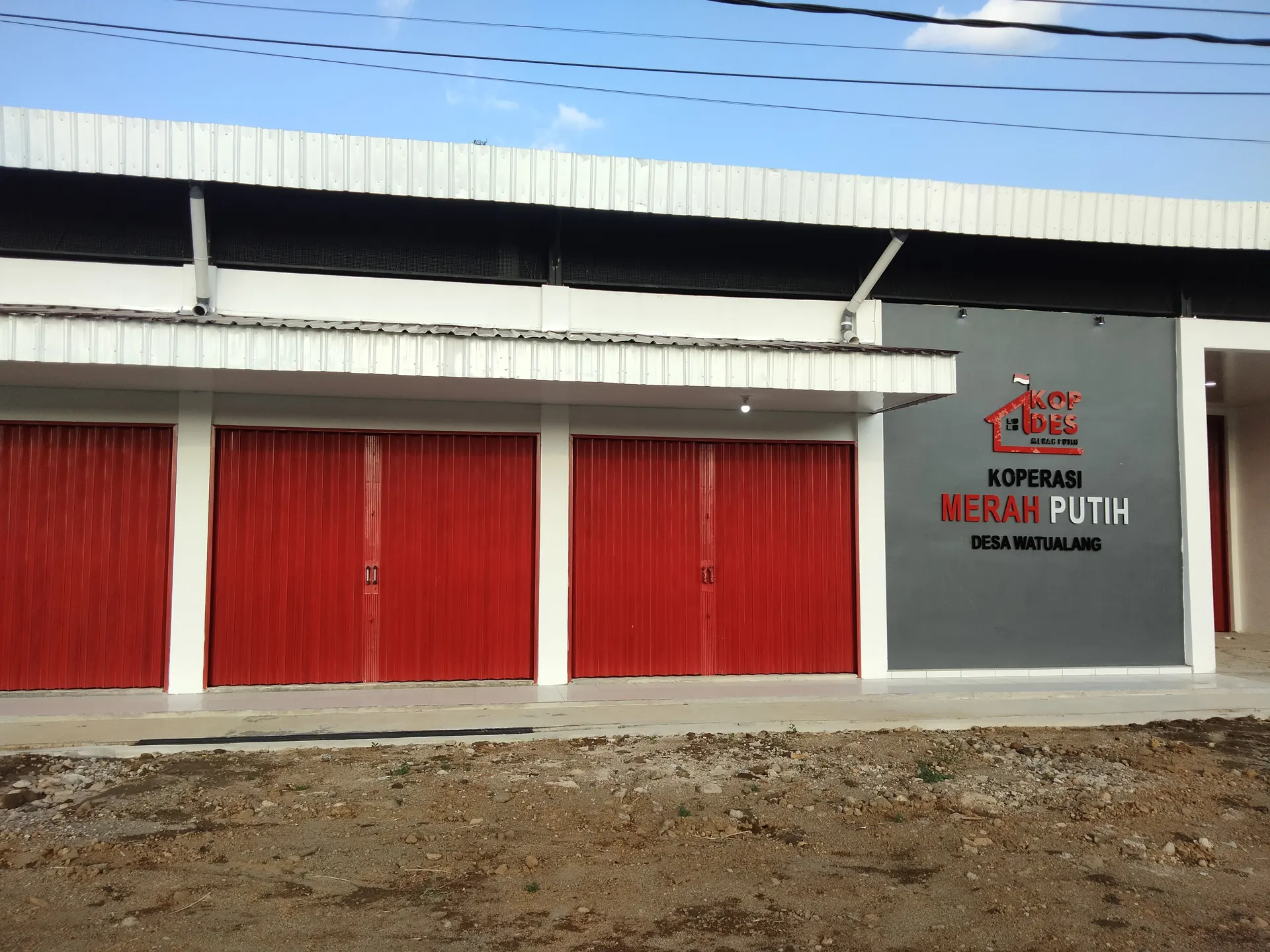 Gerai Koperasi Desa Merah Putih Desa Watualang dengan pintu rolling door merah dan papan nama Kopdes