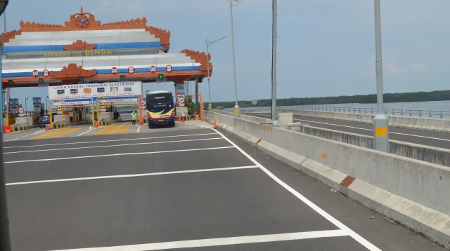 Gerbang Tol Benoa di Tol Bali Mandara dengan atap ornamen Bali, papan lajur khusus transaksi elektronik, dan sebuah bus yang melintas