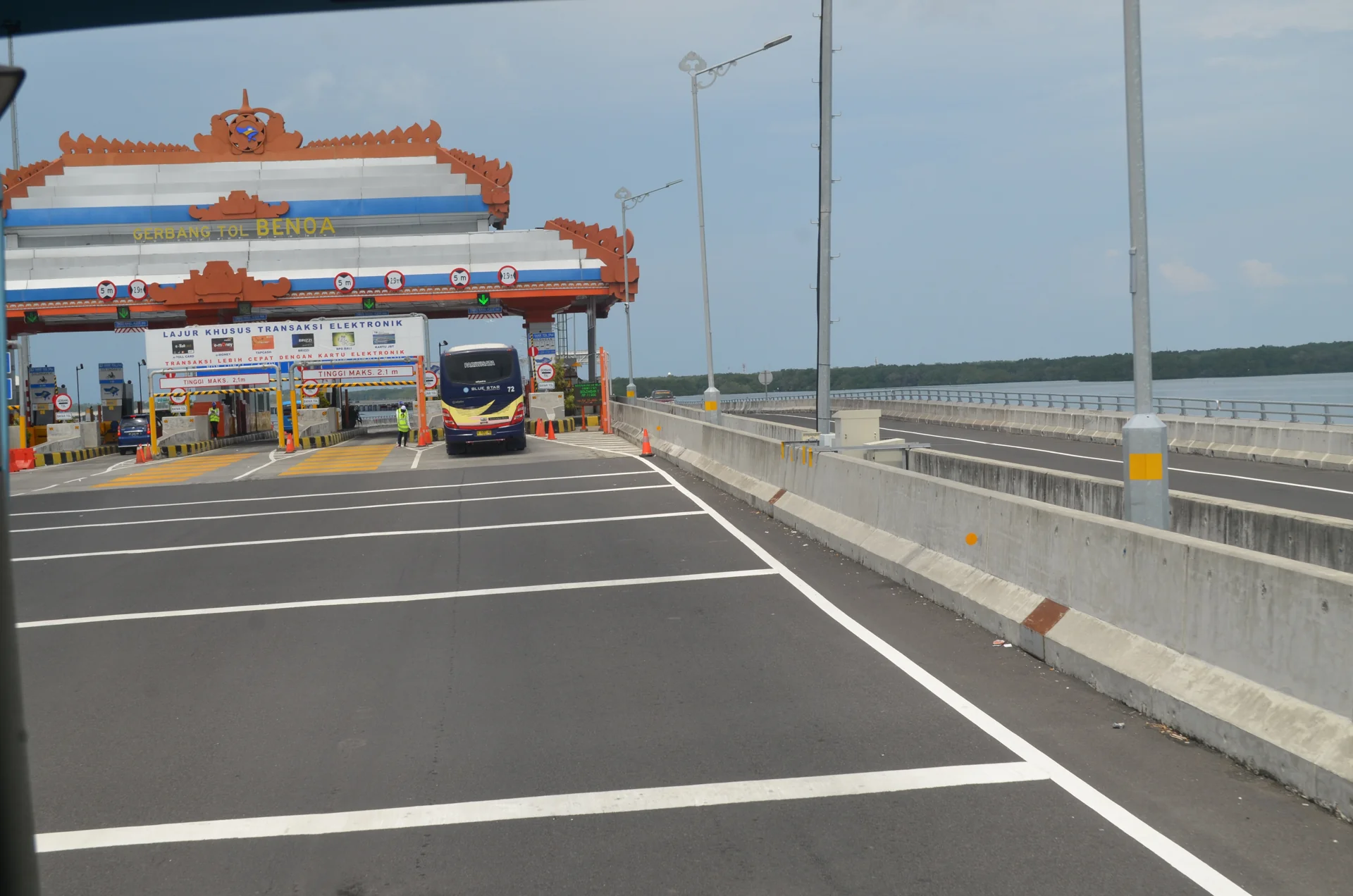Gerbang Tol Benoa di Tol Bali Mandara dengan atap ornamen Bali, papan lajur khusus transaksi elektronik, dan sebuah bus yang melintas
