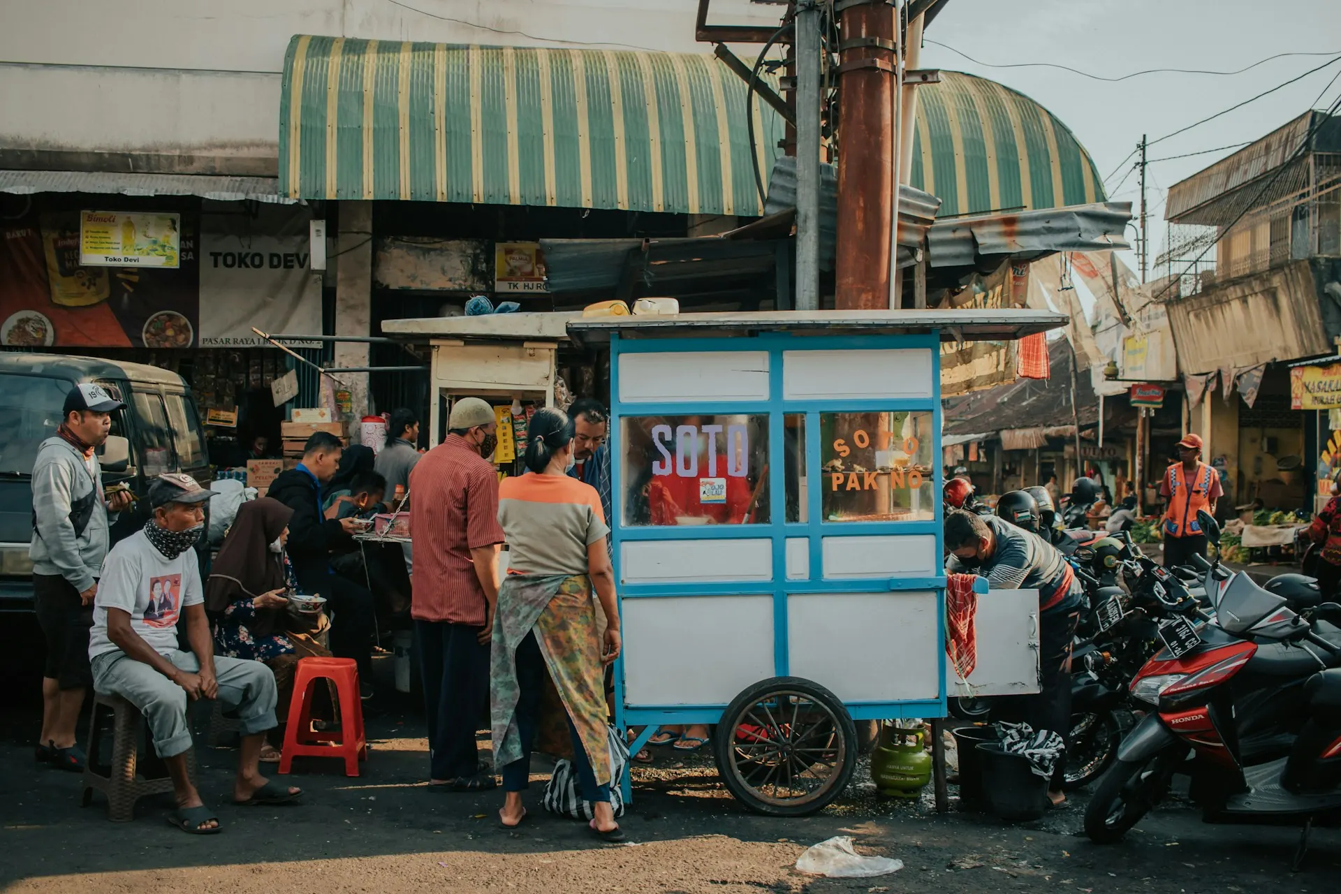 Gerobak soto biru-putih di depan deretan warung pinggir jalan dengan pembeli duduk di bangku plastik