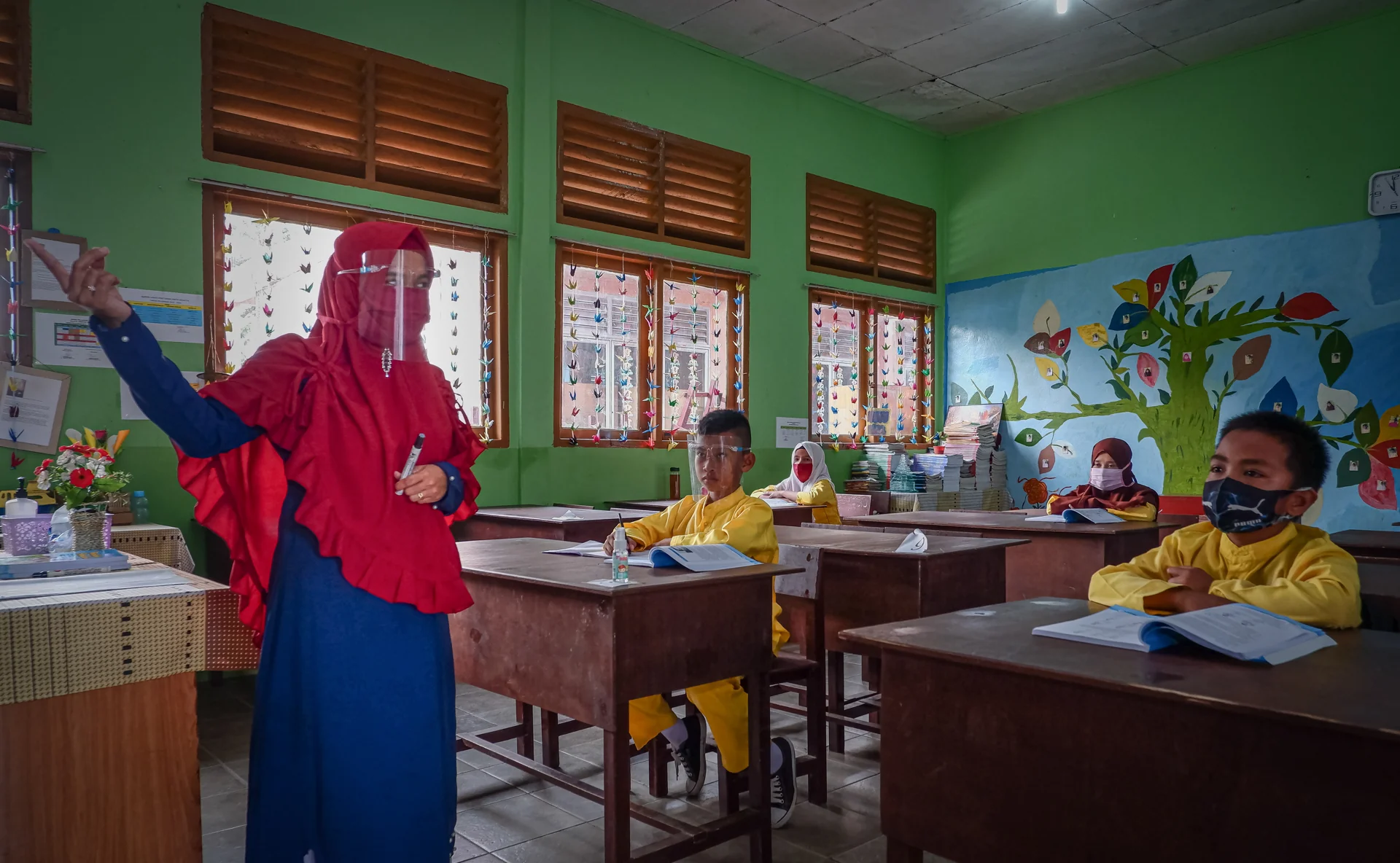 Guru berkerudung merah menerangkan pelajaran di depan kelas kepada siswa bermasker