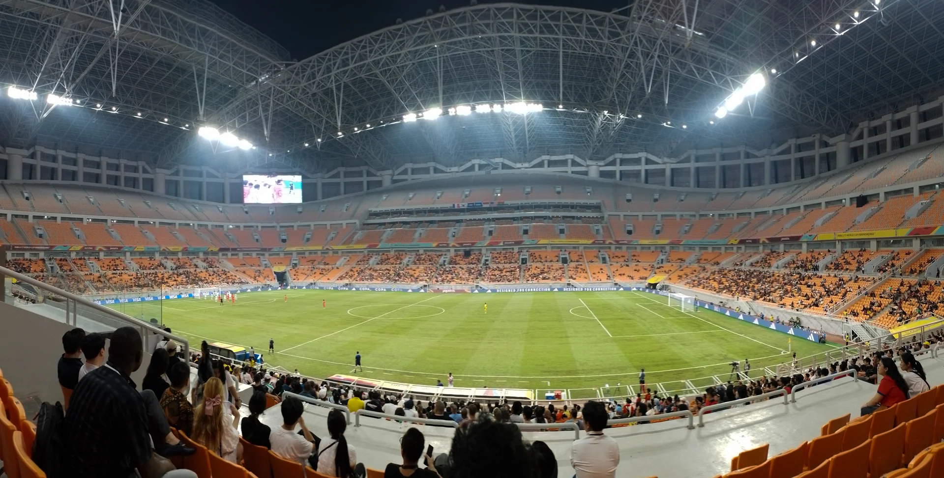 Interior Jakarta International Stadium saat pertandingan sepak bola malam hari dengan penonton di tribun