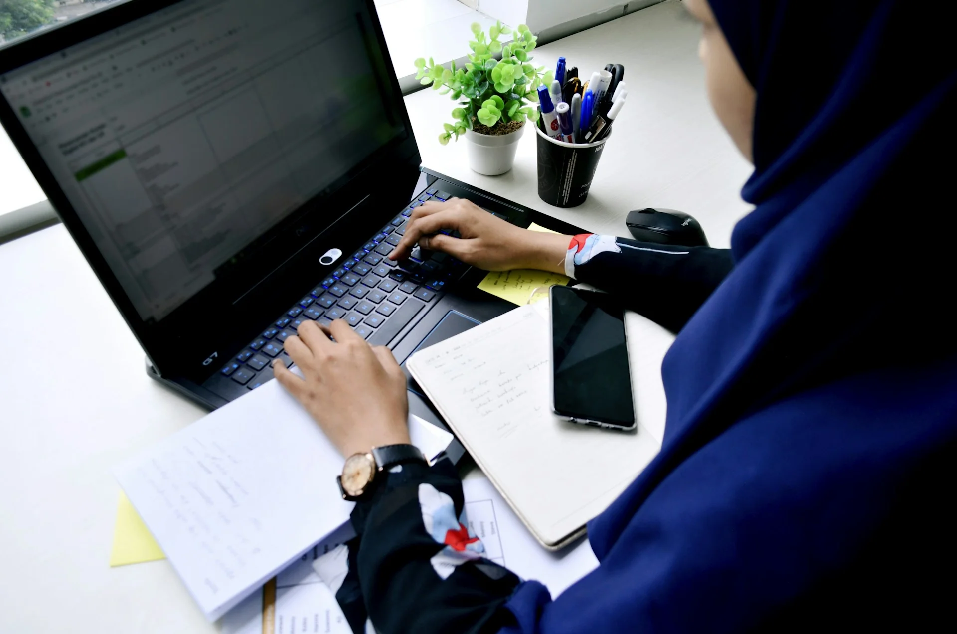 Karyawati berhijab mengetik di laptop di meja kerja dengan buku catatan dan ponsel