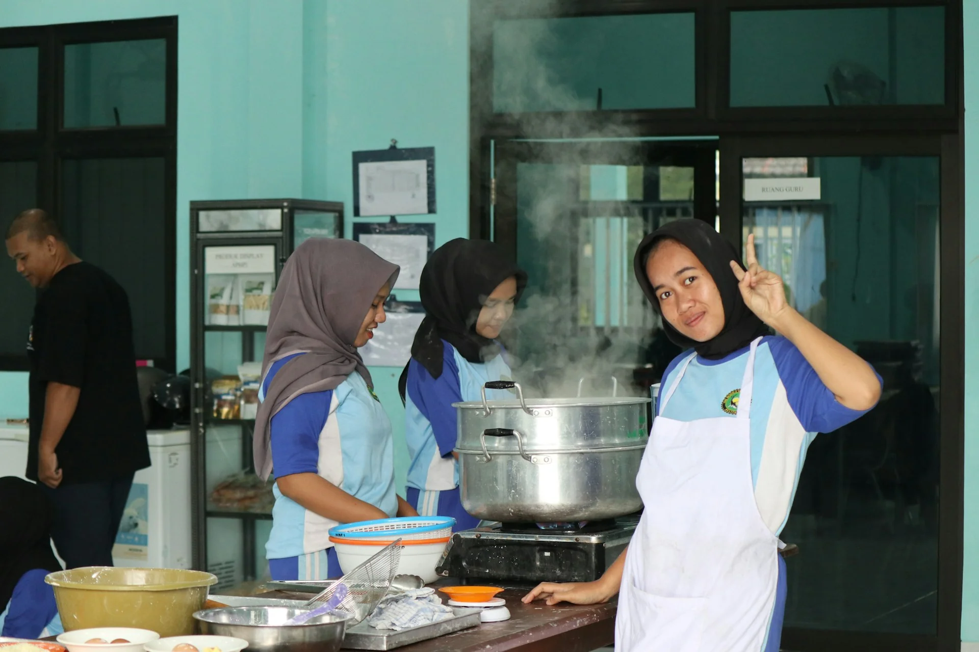 Beberapa perempuan berseragam dan celemek memasak dengan dandang kukus besar beruap di dapur