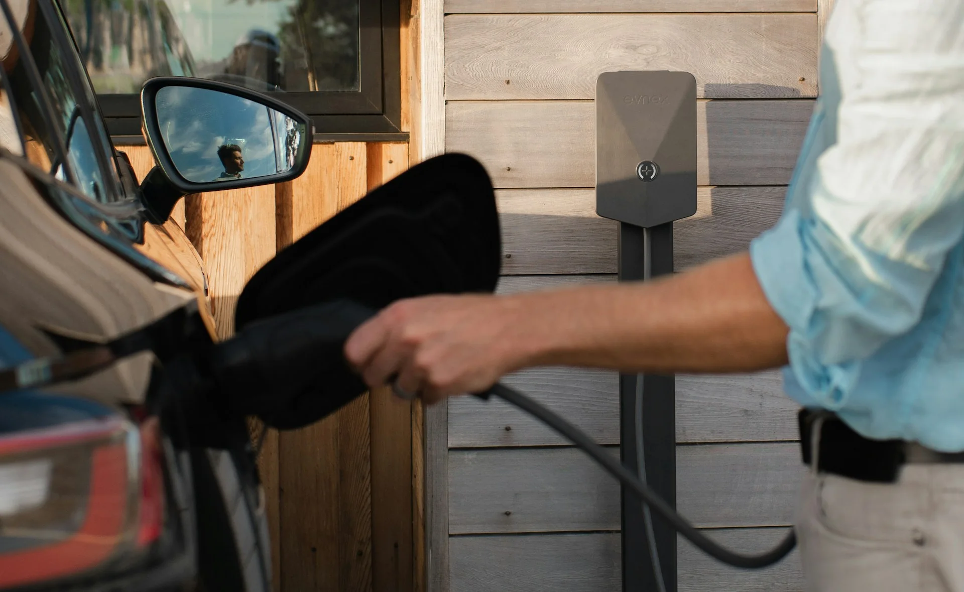 Pria mencolokkan konektor charger rumah ke port mobil listrik, dengan unit wallbox terpasang di tiang depan dinding kayu