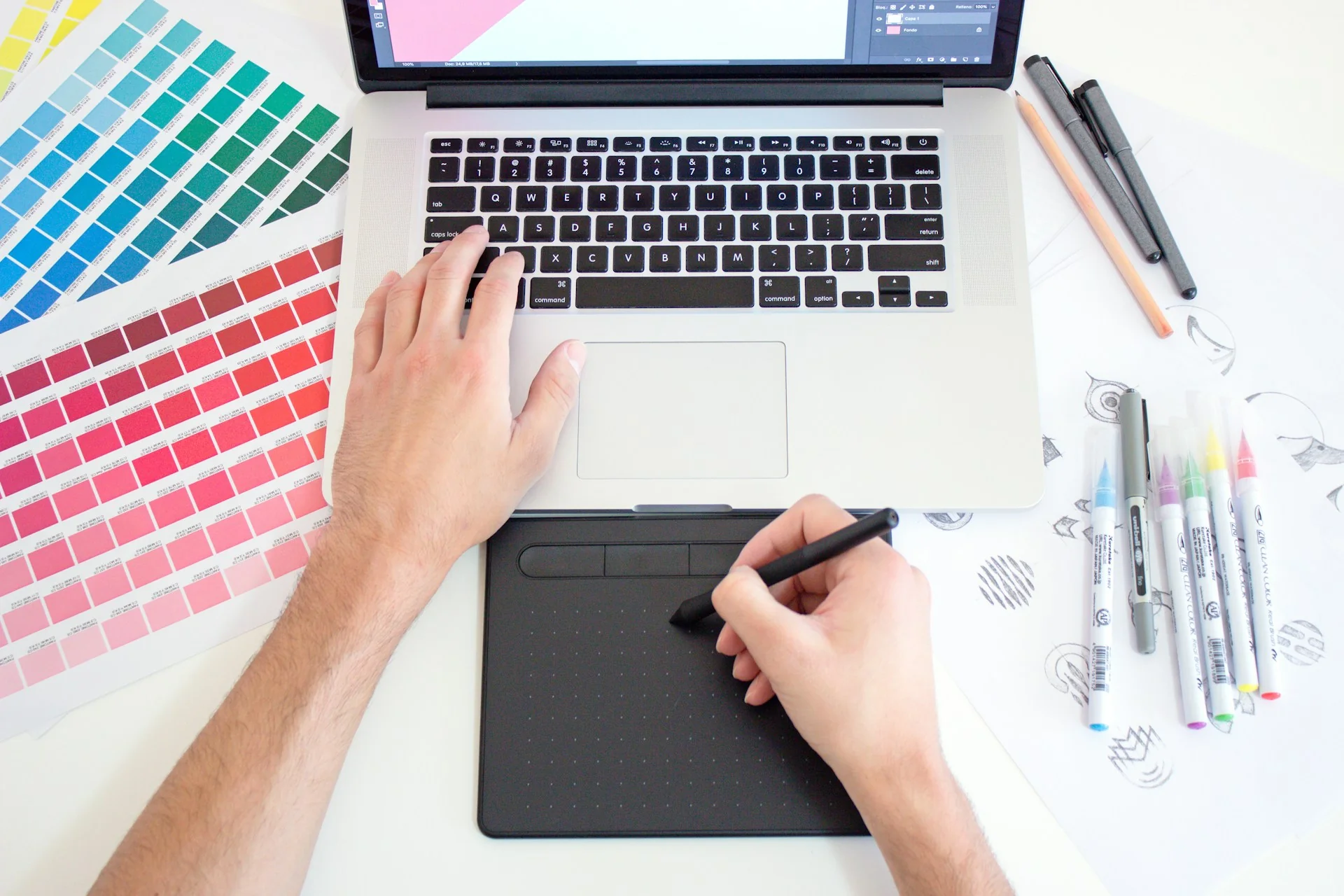 Tangan mendesain grafis dengan laptop dan pen tablet, di samping kartu palet warna dan spidol