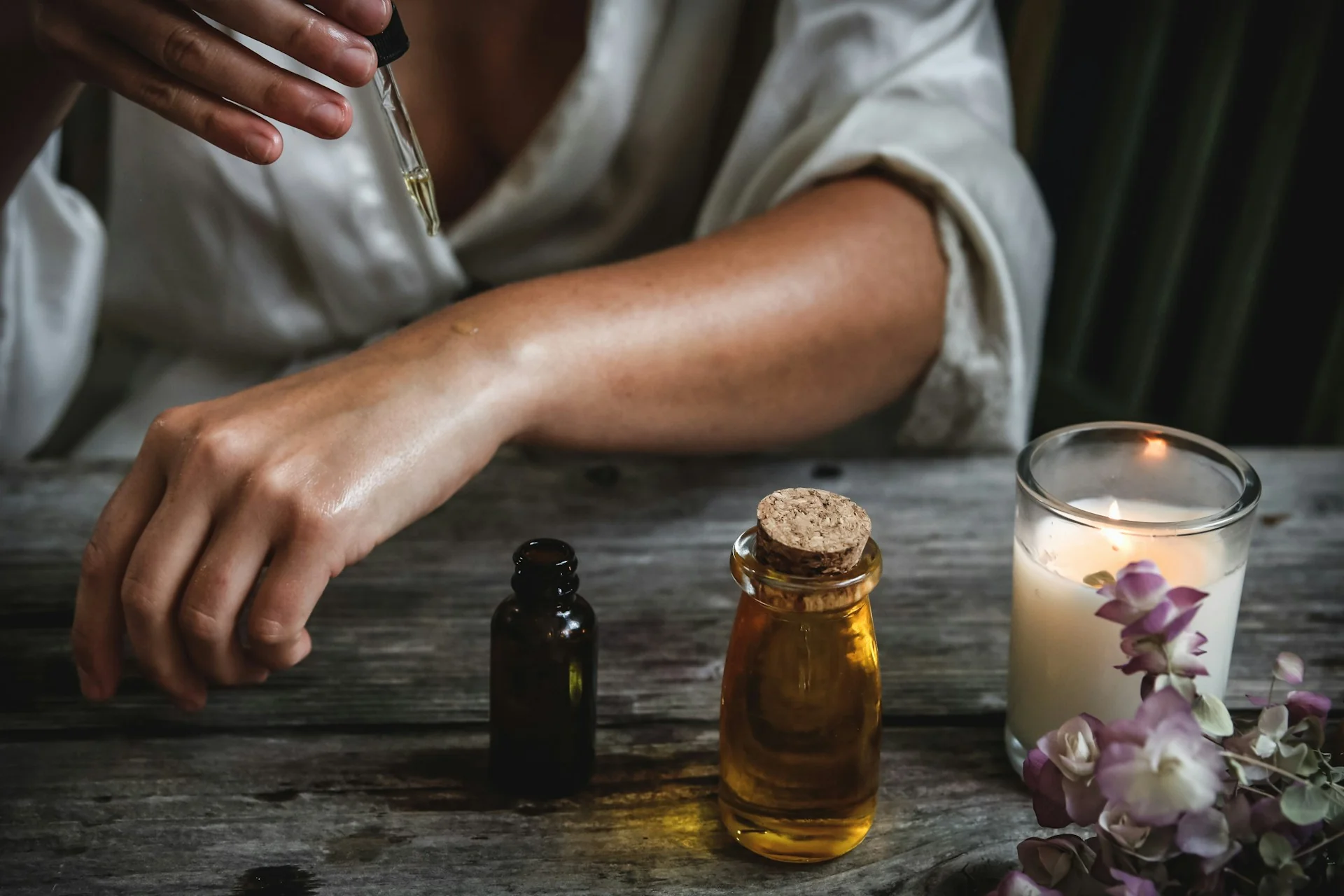 Tangan meneteskan serum dari pipet kaca ke lengan bawah, di samping botol kaca dan lilin