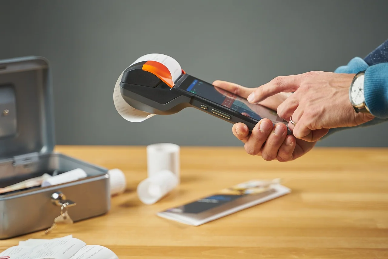 Tangan memegang mesin kasir Android genggam dengan printer thermal bawaan yang sedang mengeluarkan struk