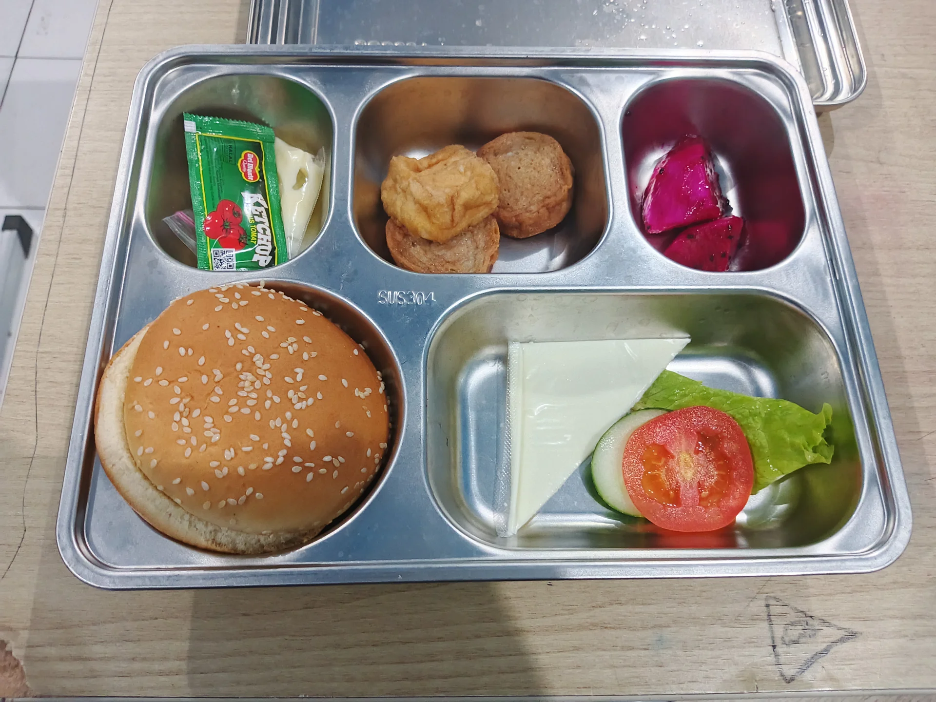 Ompreng baja tahan karat berisi roti burger, tahu goreng, buah naga, dan sayuran