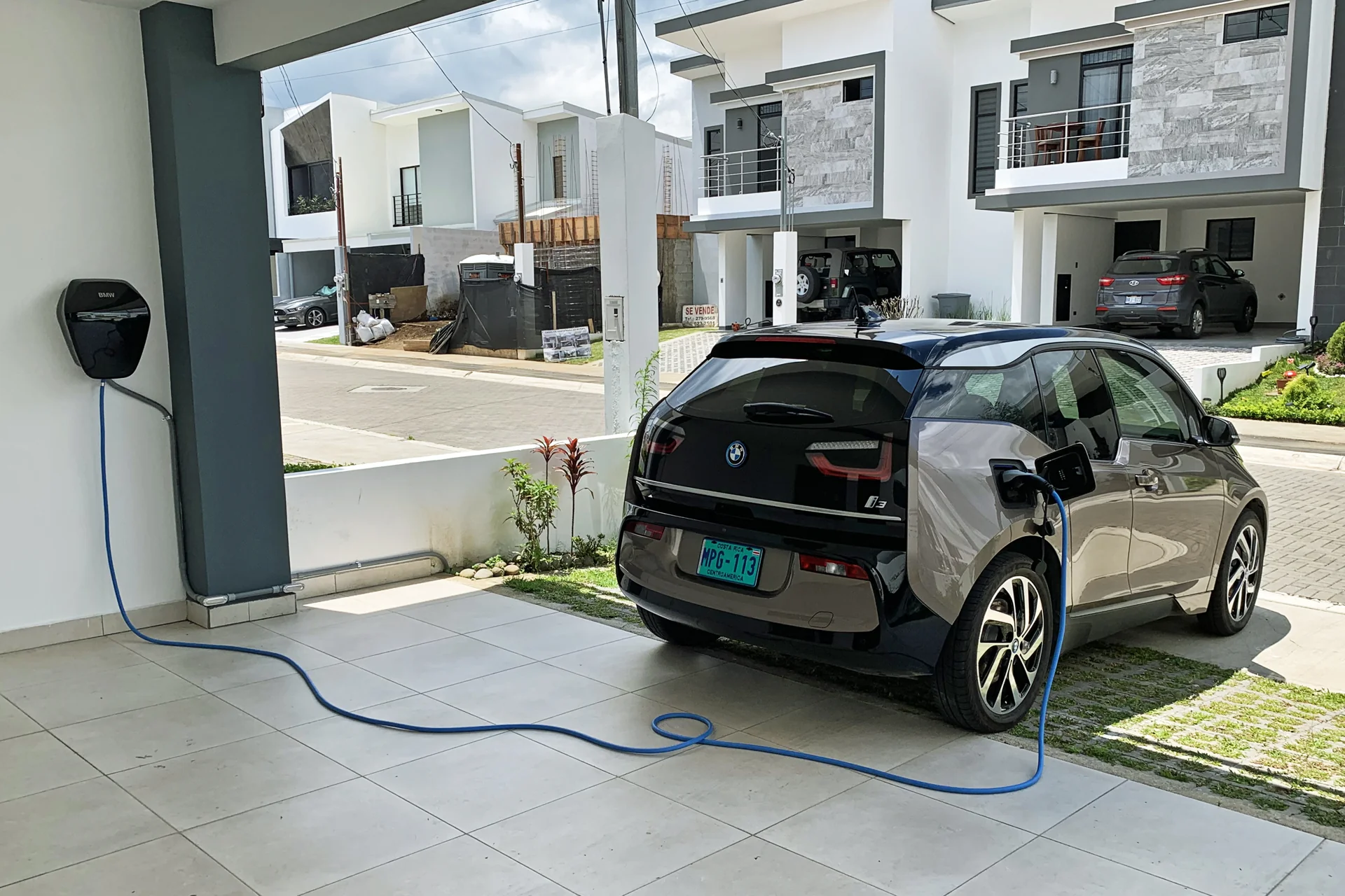 Contoh pasang charger mobil listrik di rumah: wallbox di dinding carport tersambung kabel biru ke BMW i3 yang sedang diisi