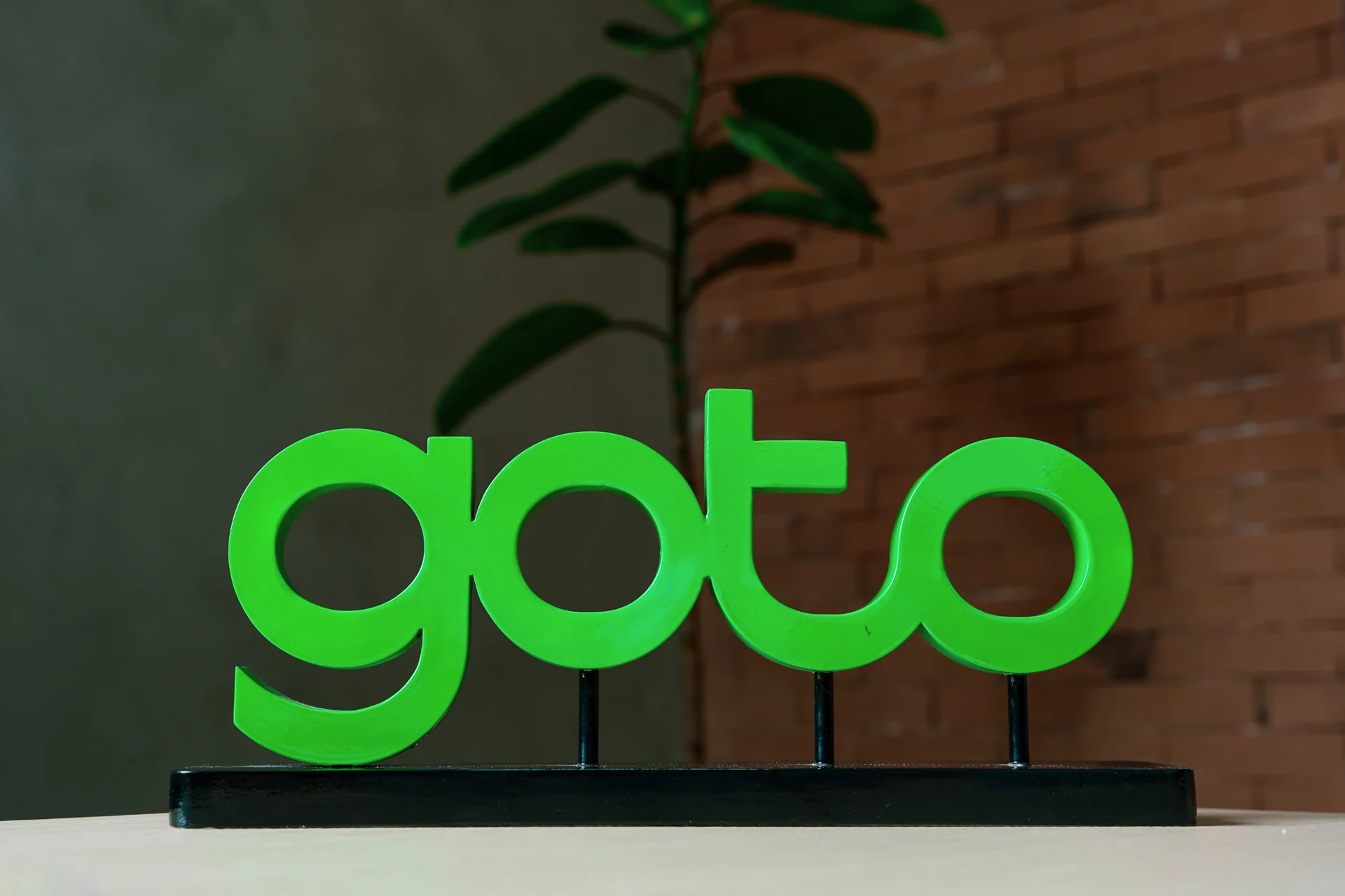 Patung logo GoTo berwarna hijau di atas meja dengan latar tanaman dan dinding bata