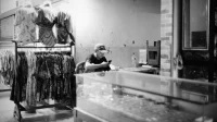 Pria bertopi memakai ponsel di meja toko pakaian batik, foto hitam putih