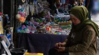 Pedagang perempuan berkerudung hijau duduk di depan kios aksesori dan perlengkapan rumah tangga di pasar