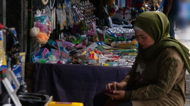 Pedagang perempuan berkerudung hijau duduk di depan kios aksesori dan perlengkapan rumah tangga di pasar