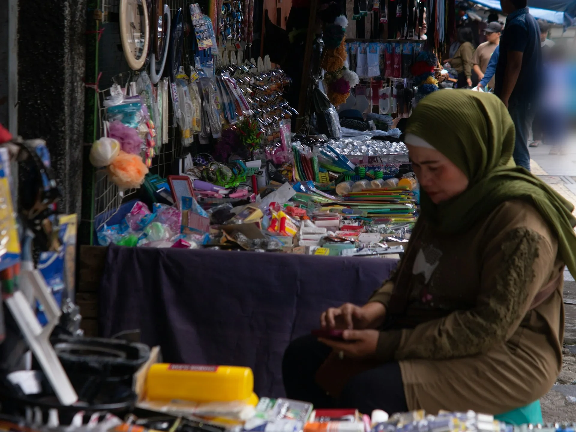 Pedagang perempuan berkerudung hijau duduk di depan kios aksesori dan perlengkapan rumah tangga di pasar