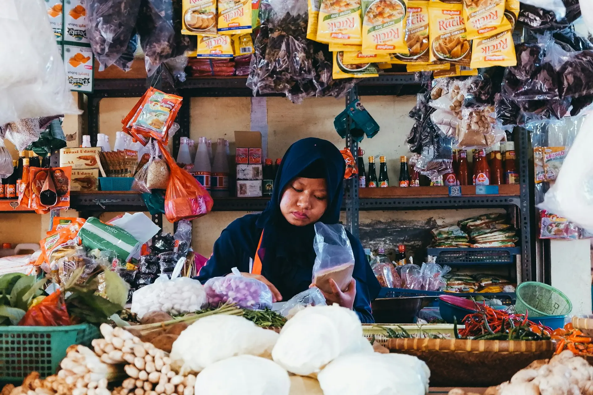 Pedagang perempuan berkerudung di kios sembako pasar dikelilingi sayuran, bumbu, dan barang kemasan