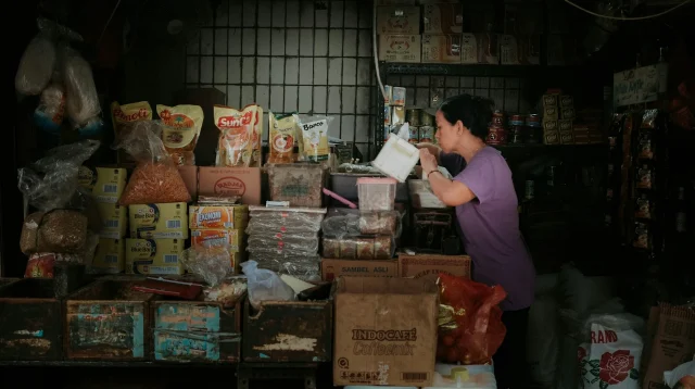 Pedagang perempuan menata barang di toko kelontong yang penuh kardus dan kemasan bahan pokok