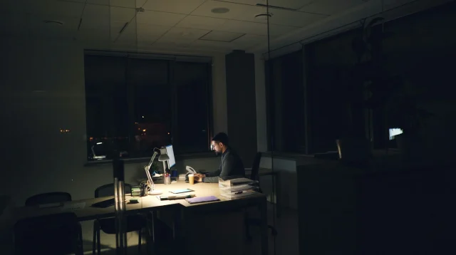 Pekerja kantor lembur malam di meja Pekerja kantor lembur sendirian di meja dengan lampu meja dan monitor di ruang kantor yang gelap