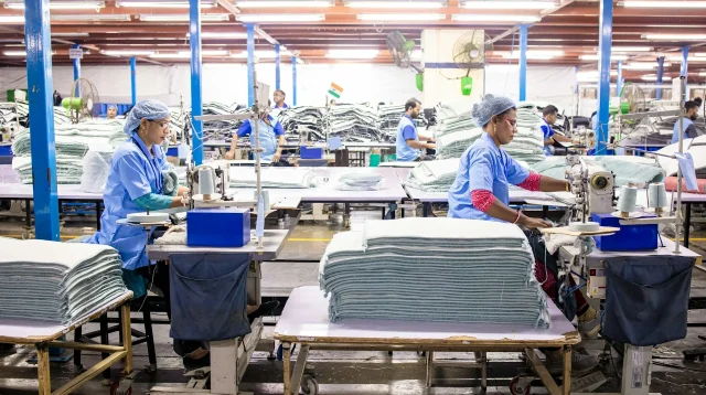 Pekerja menjahit di lini produksi pabrik tekstil Pekerja berseragam biru menjahit kain di deretan mesin jahit pabrik tekstil