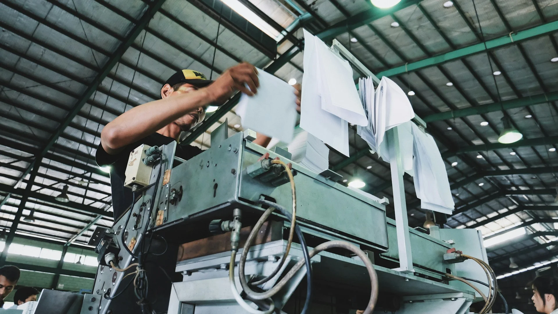 Pekerja pabrik bertopi mengoperasikan mesin cetak kertas di gudang produksi beratap baja
