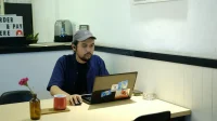Pria bertopi bekerja dengan laptop di meja kedai kopi kecil di Malang, gambaran pelaku UMKM