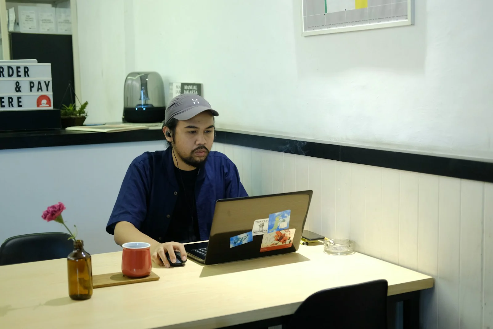 Pria bertopi bekerja dengan laptop di meja kedai kopi kecil di Malang, gambaran pelaku UMKM