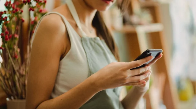 Pemilik toko berapron memegang ponsel dan mengetik balasan chat pelanggan di dalam tokonya