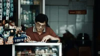 Pemilik toko berkacamata mencatat di kertas dengan kalkulator di meja etalase tokonya