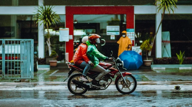 Dua pengendara motor berjas hujan merah dan hijau melintas di jalan basah saat hujan di Malang