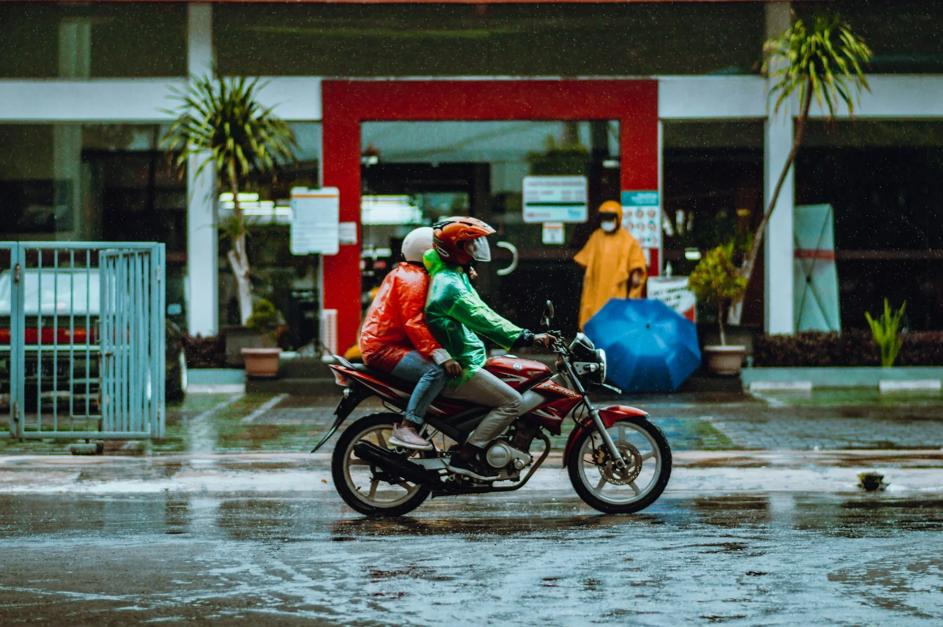 Dua pengendara motor berjas hujan merah dan hijau melintas di jalan basah saat hujan di Malang
