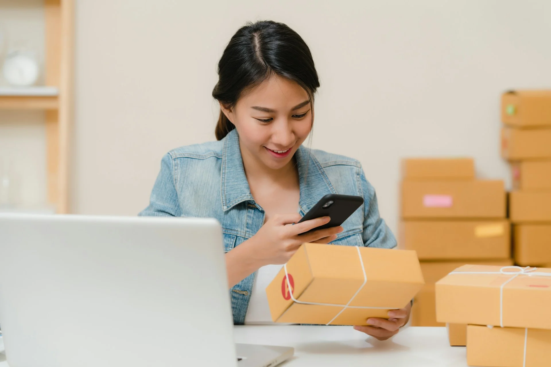 Penjual daring memotret paket pesanan dengan ponsel di samping laptop dan tumpukan kardus