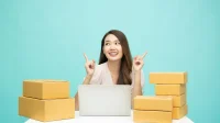 Perempuan pemilik toko online duduk di depan laptop di antara tumpukan kardus paket pesanan