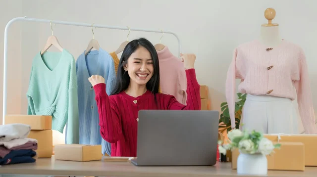 Penjual pakaian online mengepalkan tangan gembira di depan laptop, dikelilingi rak baju, manekin, dan kardus pesanan