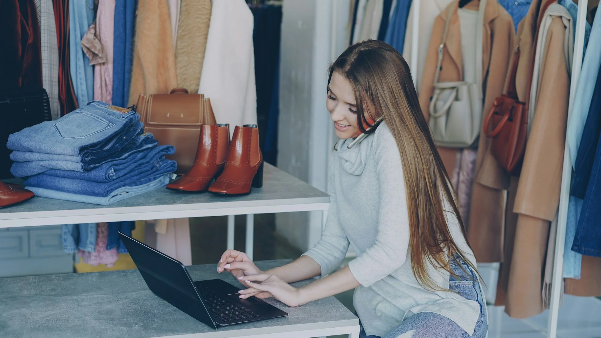 Pemilik toko pakaian menelepon sambil memeriksa laptop di samping tumpukan celana jeans dan sepatu bot