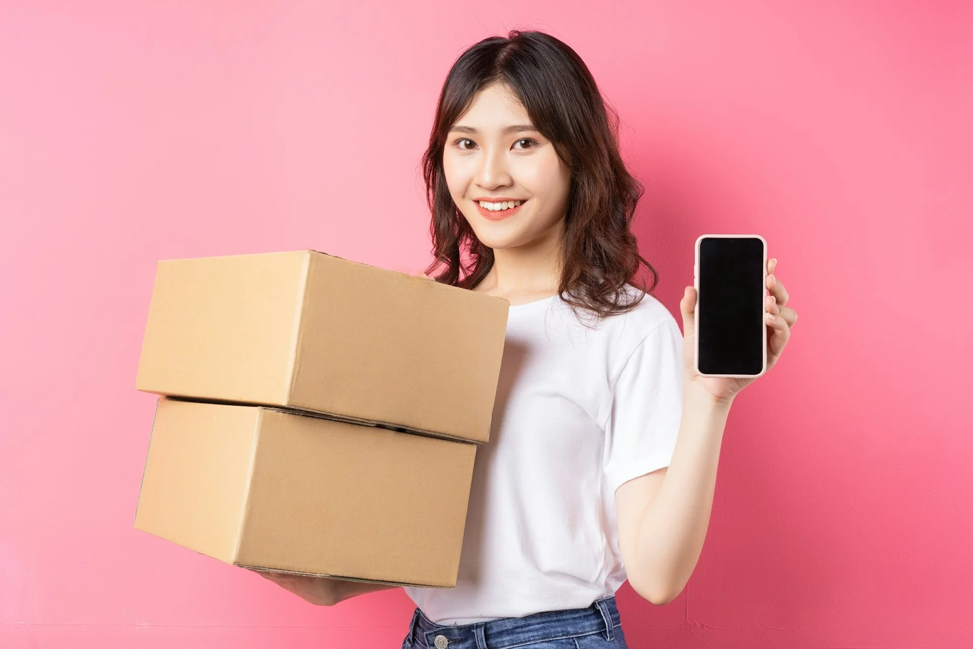 Perempuan berkaus putih memegang dua kardus paket dan menunjukkan ponsel