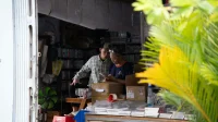 Dua pemilik toko kecil memeriksa pesanan di ponsel di antara kardus paket dan tumpukan barang