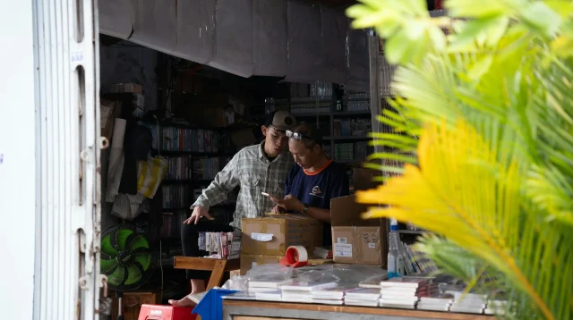 Dua pemilik toko kecil memeriksa pesanan di ponsel di antara kardus paket dan tumpukan barang