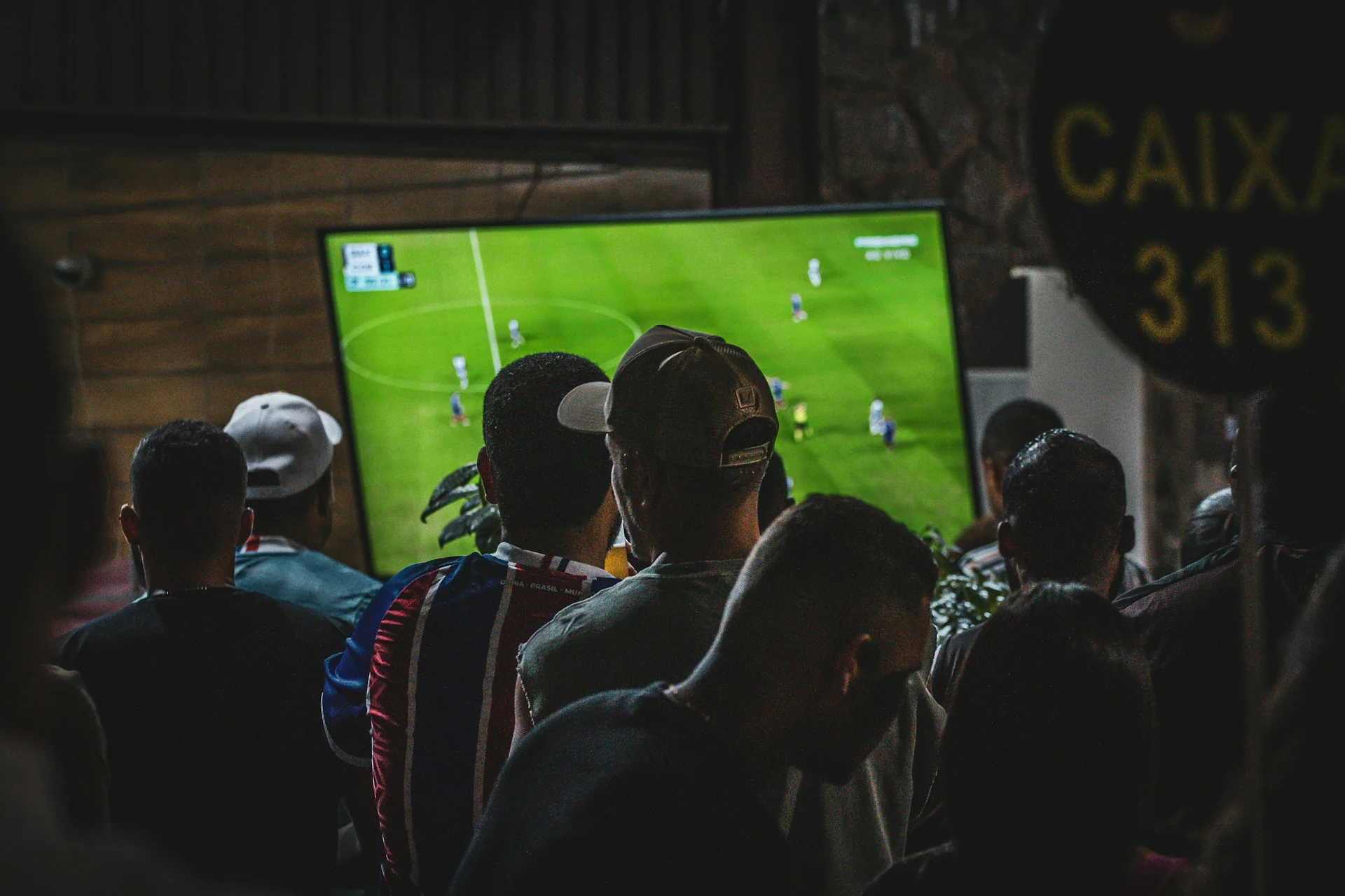 Sekelompok penonton menyaksikan siaran pertandingan sepak bola di layar televisi besar dalam ruangan