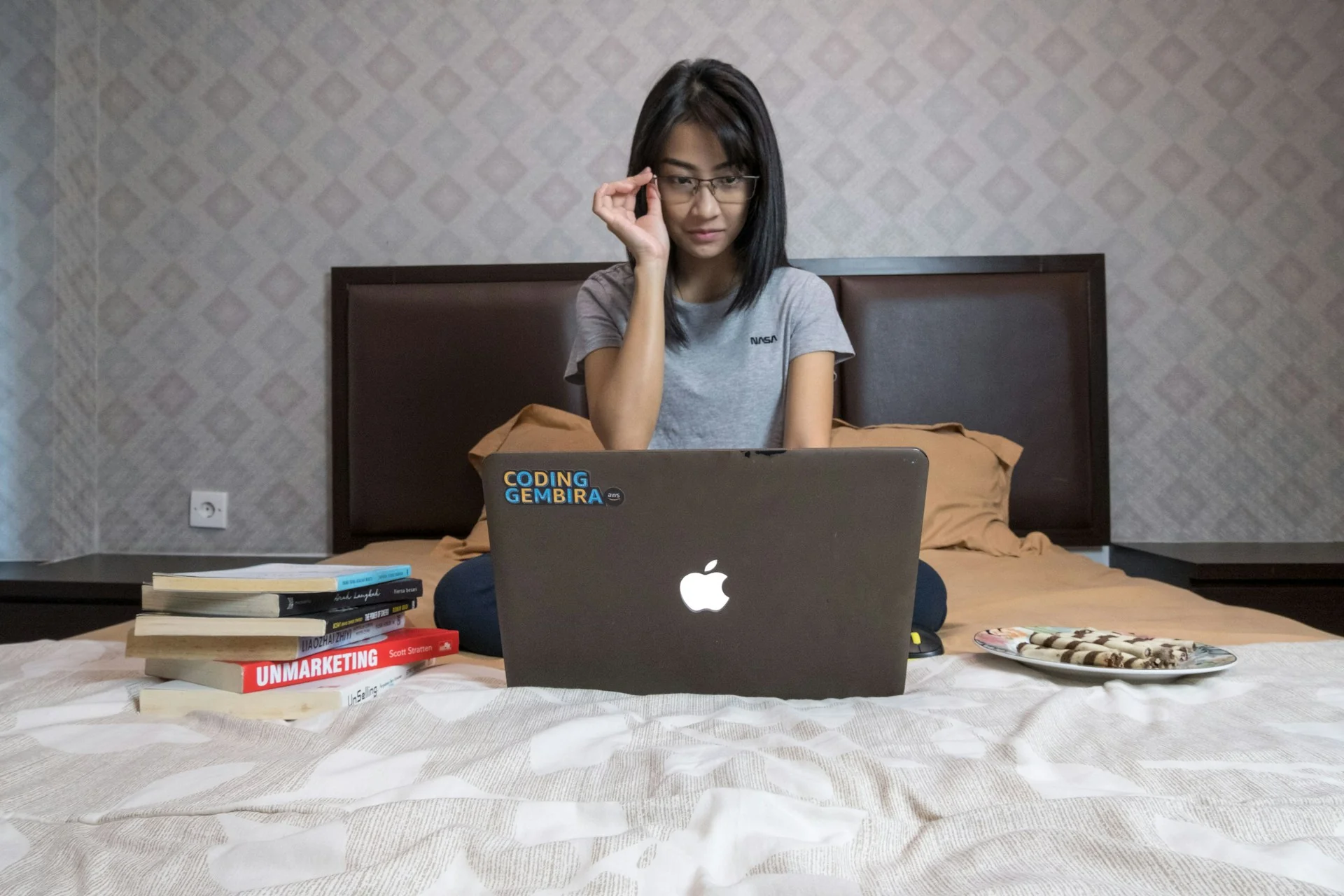 Perempuan berkacamata duduk di atas tempat tidur menatap layar laptop, ditemani tumpukan buku dan sepiring kue