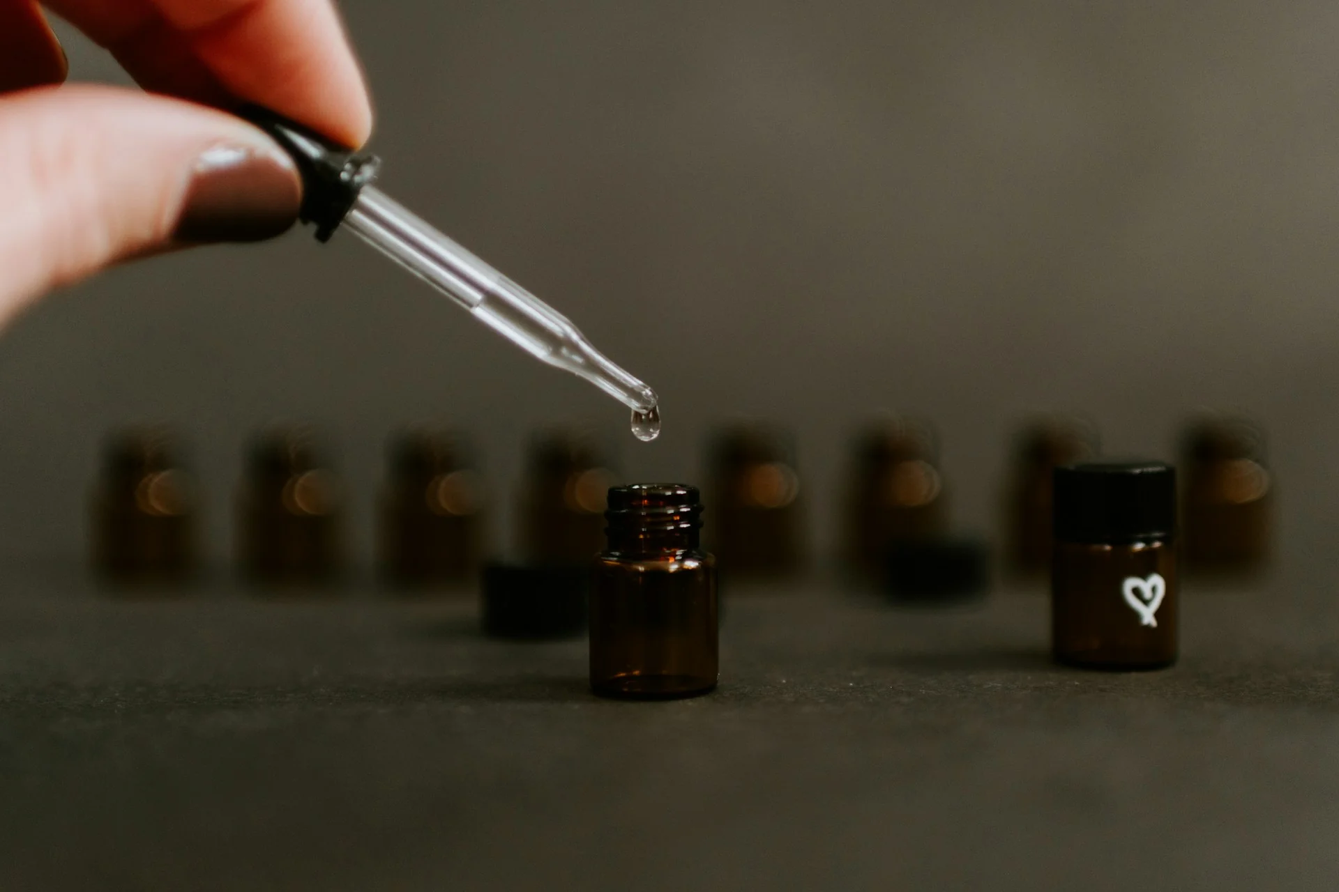 Tangan memegang pipet kaca yang meneteskan satu tetes cairan ke botol kaca amber kecil
