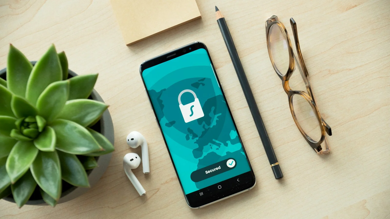 Ponsel di meja kayu menampilkan ikon gembok dan tulisan Secured, di samping earbud, pensil, dan kacamata
