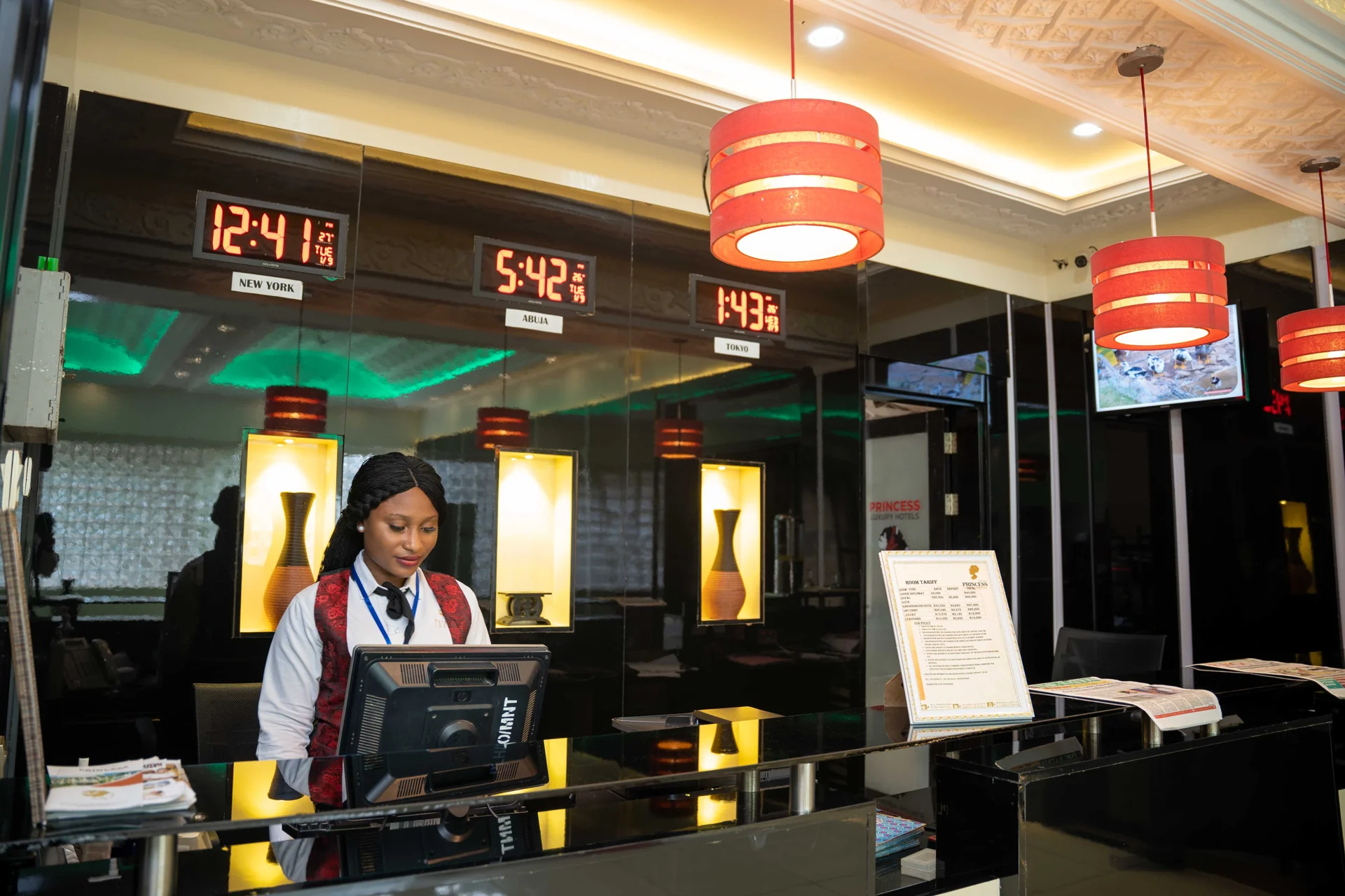 Resepsionis hotel berdiri di balik meja depan dengan monitor dan jam dinding beberapa kota