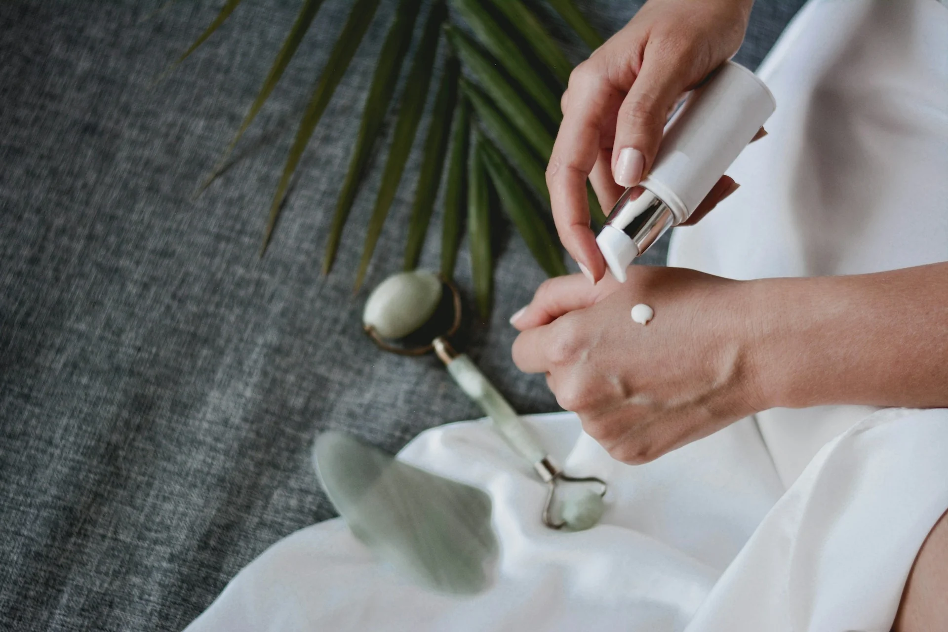 Tangan meneteskan sedikit produk perawatan kulit dari botol putih ke punggung tangan, dengan jade roller di latar