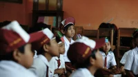 Siswa sekolah dasar berseragam putih dengan topi dan dasi merah duduk di lantai kelas