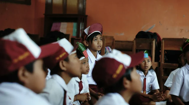 Siswa sekolah dasar berseragam putih dengan topi dan dasi merah duduk di lantai kelas